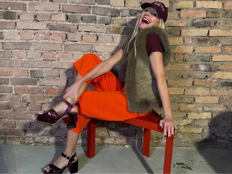 Lachende Renate Zott in weiten orangen Hosen, Fellweste und Samtsandalen posiert lässig vor alter Backsteinmauer.