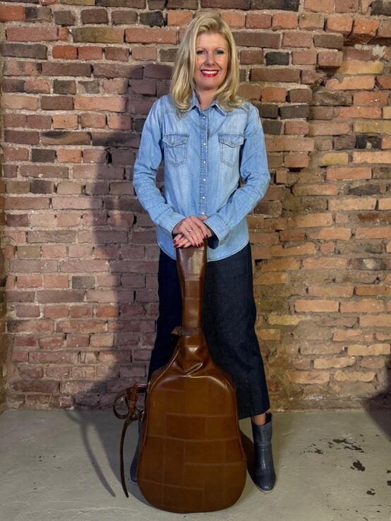 Lachende Renate Zott mittleren Alters mit offenem Haar, Jeanshemd und Leder-Gitarrentasche vor alter Backsteinwand. | © Renate Zott