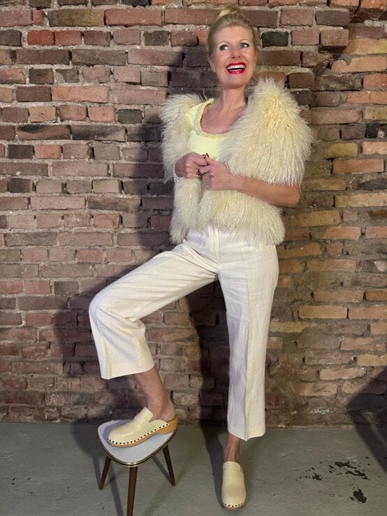 Blonde Renate Zott mittleren Alters in heller Marlenehose, flauschiger Fellweste und hellen Pantoletten lächelt vor Ziegelwand. | © Renate Zott