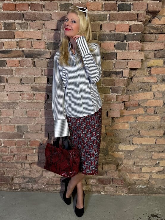 Blonde Renate Zott mit rotgemustertem Rock, gestreifter Bluse und roter Handtasche lehnt lächelnd vor Backsteinmauer. | © Renate Zott