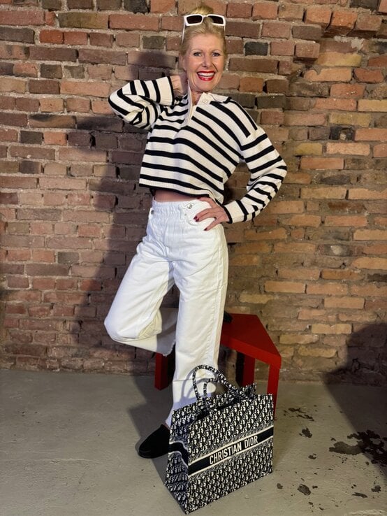 Lachende Renate Zott mit weiß-schwarz gestreiftem Pullover, weißer Hose und großer Dior-Tasche vor Backsteinmauer. | © Renate Zott