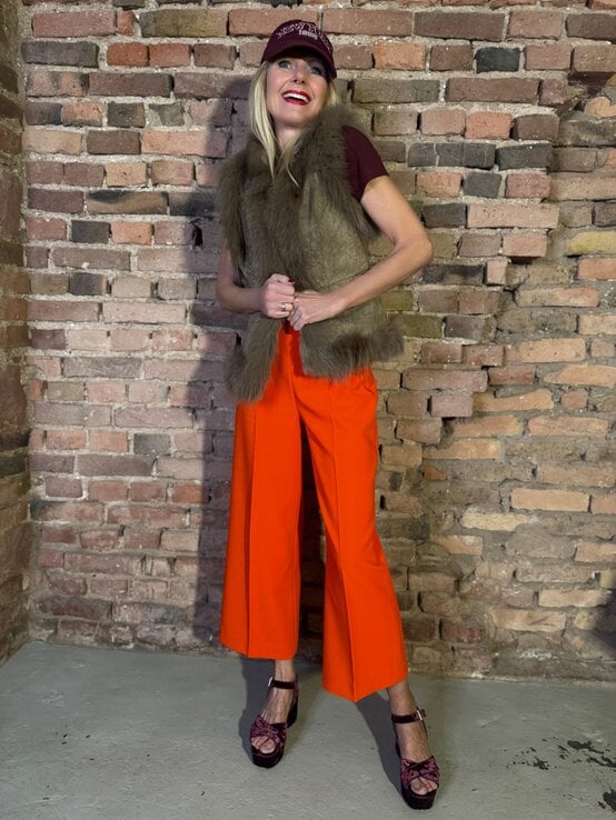 Lachende Renate Zott mit bordeauxfarbener Kappe, Fellweste und orangefarbener Hose steht vor Backsteinwand. | © Renate Zott