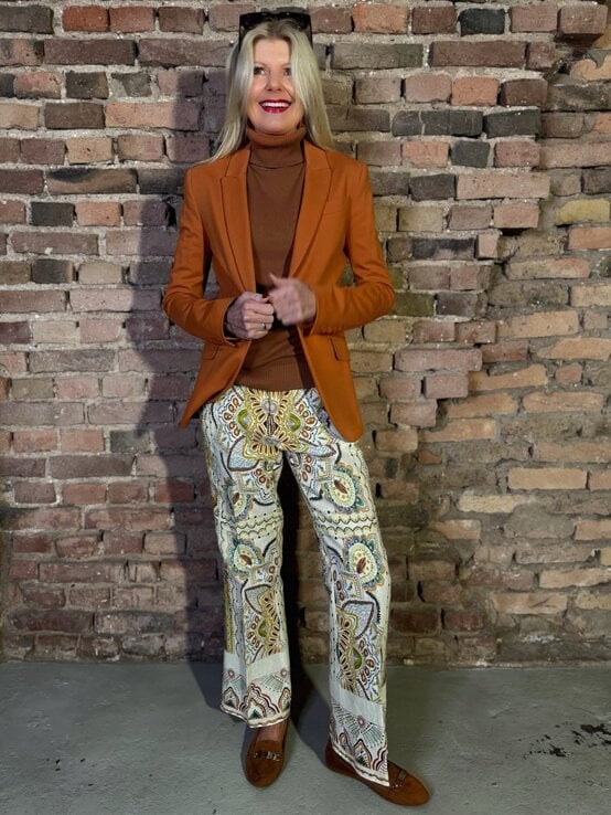 Renate Zott mit braunem Rollkragen, orangefarbenem Blazer und gemusterten Schlaghosen lächelt vor Backsteinwand. | © Renate Zott