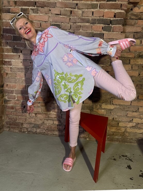 Renate Zott in pastellfarbenem Outfit mit floralem Muster steht lachend und locker vor rustikaler Ziegelmauer. | © Renate Zott Frühlings