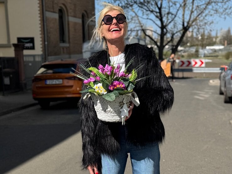 Lachende blonde Renate Zott mit schwarzer Fransjacke und Sonnenbrille hält Frühlingsblumenkorb auf sonniger Straße vor Stadtkulisse.