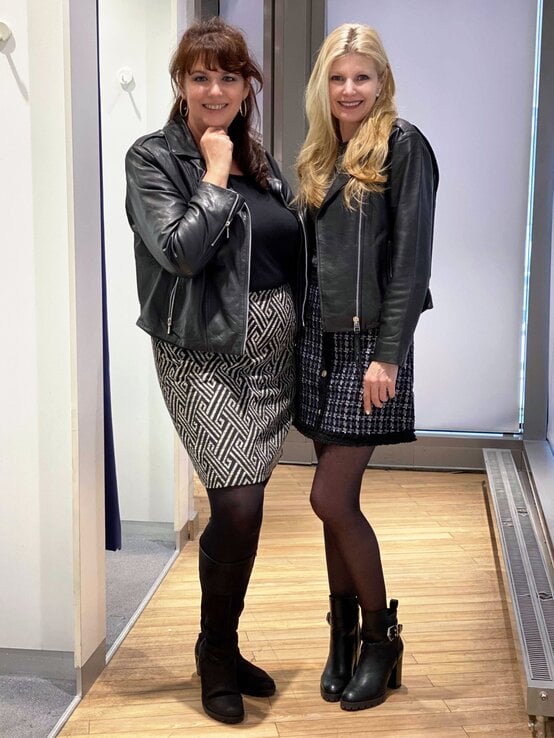 Zwei elegant gekleidete Frauen in kurzen Röcken und Lederjacken posieren lächelnd in einem hellen Umkleideraum. | © Renate Zott