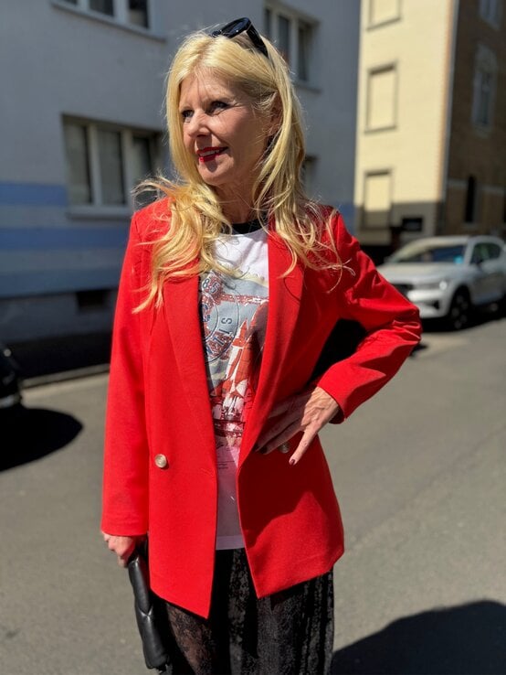 Renate Zott mit langen blonden Haaren trägt roten Blazer über Printshirt und schwarzem Spitzenrock, steht lächelnd in der Sonne. | © Renate Zott