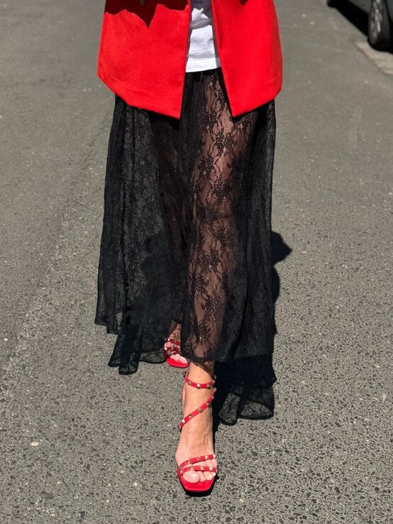 Schwarzer transparenter Spitzenrock schwingt über roten Sandaletten mit Nietenriemen auf grauem Asphalt im Sonnenlicht. | © Renate Zott