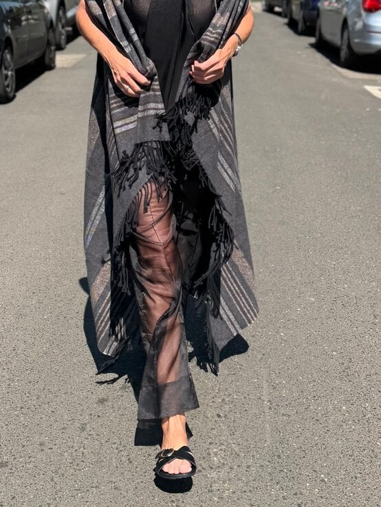 Schwarze transparente Hose unter grauem Fransencape mit Streifenmuster, kombiniert mit offenen Sandalen auf Asphalt bei Sonnenschein. | © Renate Zott