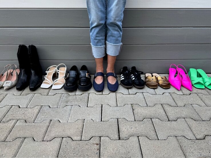 Renate Zott Eine Person in hochgekrempelter Jeans steht barfuß vor einer Wand, umgeben von zehn Paar verschiedenfarbigen Damenschuhen.