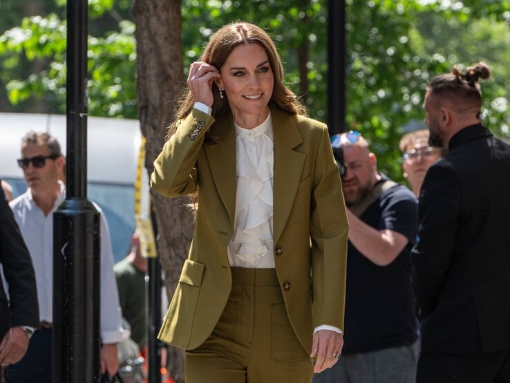 Prinzessin Kate  in olivgrünem Hosenanzug, Wedges Sandalen und weißer Rüschenbluse lächelt in die Kamera, umgeben von Fotografen und Passanten.