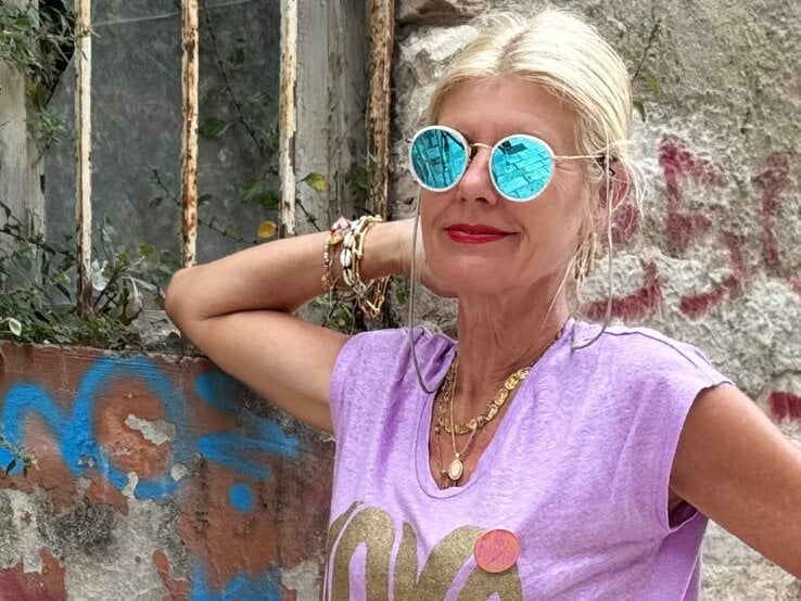 Renate Zott mit türkis verspiegelter Sonnenbrille, lilafarbenem Shirt und goldenen Ketten steht entspannt vor Graffitiwand.