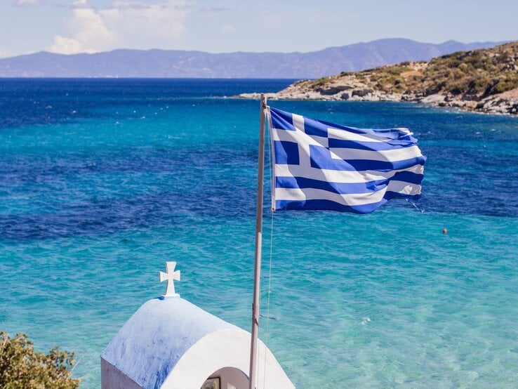 Symbolbild für Luxus-Sandalen: Die griechische Flagge weht im Wind vor türkisblauem Meer, dahinter eine kleine weiße Kapelle mit Kreuz und felsiger Küste.