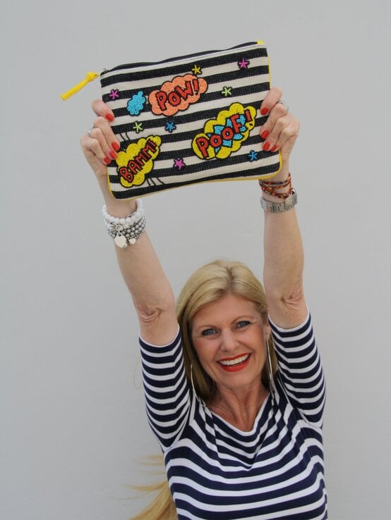 Lachende blonde Renate Zott in Ringelshirt hebt stolz eine gestreifte Clutch mit Comic-Schriftzügen wie „POW!“ und „BAMM!“ in die Höhe. | © Renate Zott 