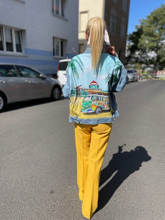 Renate Zott mit hohem Zopf und Haarband geht in gelber Schlaghose und bunter Jacke mit Strandmotiv eine sonnige Straße entlang. | © Renate Zott 