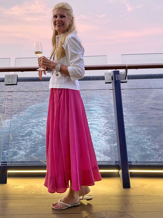 Renate Zott im weißen Blazer und pinkem Maxirock lächelt mit Weinglas auf dem Deck eines Schiffs bei rosa Abendhimmel. | © Renate Zott 