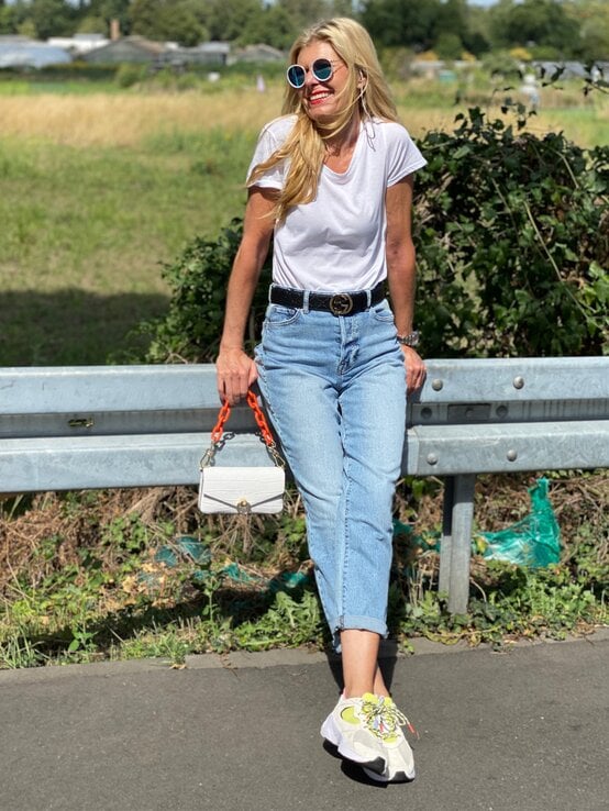 Fröhliche Renate Zott mit weißem T-Shirt und heller Jeans lehnt entspannt an Leitplanke, trägt bunte Sneaker und Sonnenbrille. | © Renate Zott 