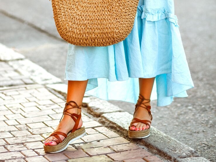 Frau mit lackierten Zehen in braunen Bast-Wedges und hellblauem Kleid steht auf Pflastersteinen mit großer Strohtasche.