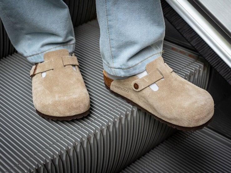 Zwei Füße in beigen Wildleder-Clogs und weißen Socken stehen auf einer fahrenden Rolltreppe aus Metallrillen.