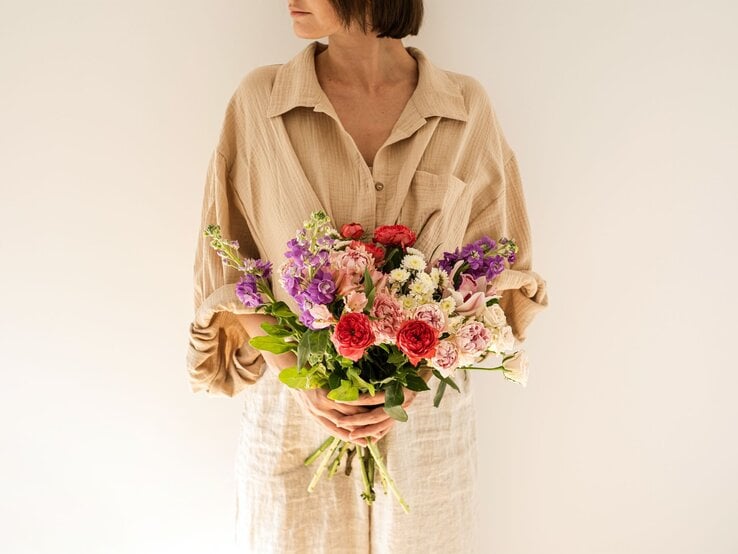 Frau mit kurzem braunem Haar hält einen bunten Strauß Blumen vor cremefarbenem Hintergrund und blickt zur Seite.
