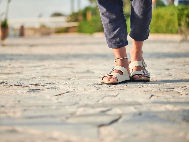 Füße in beigen Birkenstock-Sandalen und hochgekrempelter Hose auf heller Natursteinpromenade bei warmem Abendlicht.