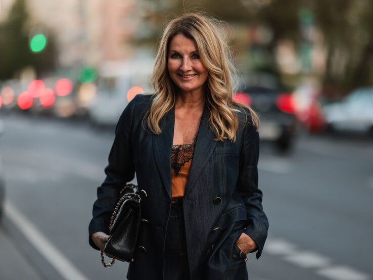 Frauke Ludowig  mit schwarzem Blazer und Handtasche lächelt selbstbewusst auf einer belebten Straße.