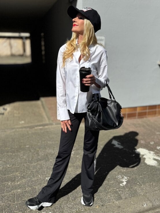 Renate Zott in weißem Hemd, schwarzer Kappe und Sneakers steht mit Kaffeebecher lächelnd im Sonnenlicht auf Asphalt. | © Renate Zott 