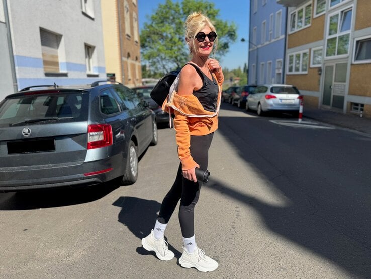 Renate Zott mit Sonnenbrille, orangefarbener Jacke und schwarzem Sportoutfit läuft lachend mit Rucksack durch Wohnstraße.
