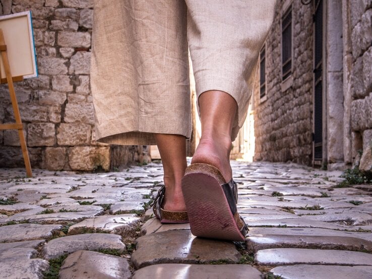 Nahaufnahme von Beinen in beigen Leinenhosen und Sandalen, die über glänzende Kopfsteinpflaster in einer engen Altstadtgasse laufen.