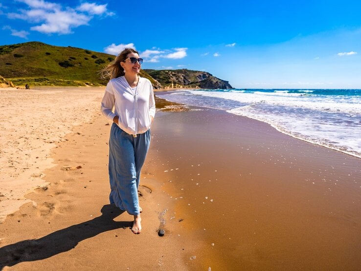 Eine Frau in weißer Bluse und blauer Hose spaziert barfuß am sonnigen Sandstrand entlang, während kleine Wellen ans Ufer schlagen.