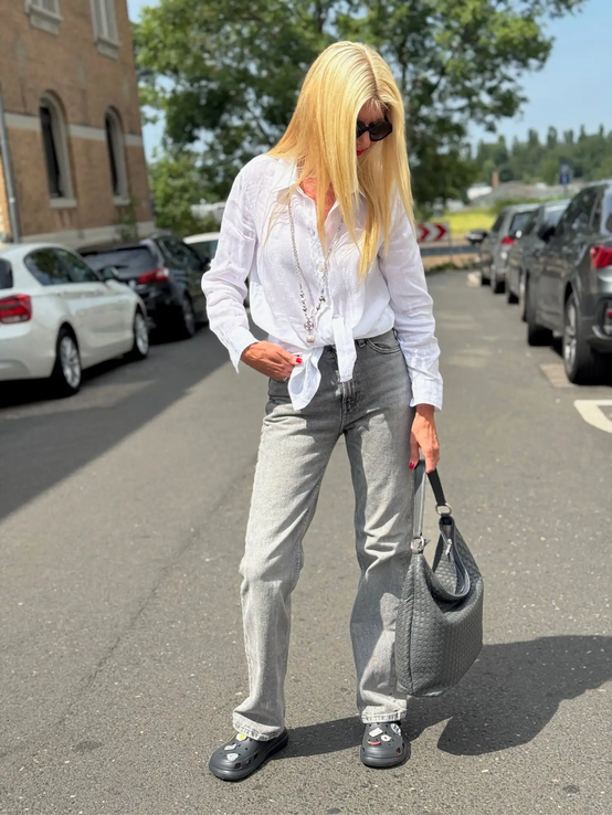 Blondierte Frau mit weißer Bluse und grauer Jeans blickt nach unten und trägt eine große schwarze Tasche auf sonniger Straße. | © Renate Zott