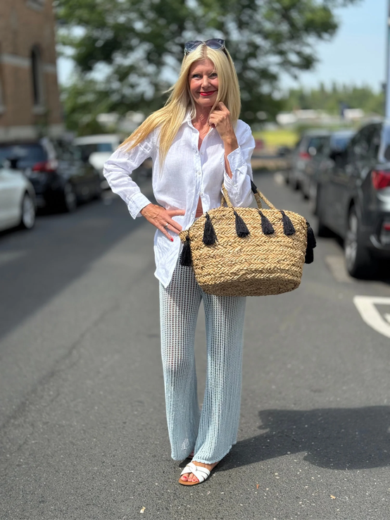 Blondierte Frau mit weißer Bluse und Häkelpants trägt lächelnd eine große Strohtasche mit schwarzen Quasten auf Straße. | © Renate Zott