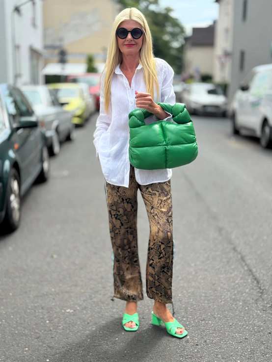 Blondierte Frau mit weißer Bluse, Schlangenprint-Hose und grüner Handtasche steht modisch gestylt auf leerer Straße. | © Renate Zott