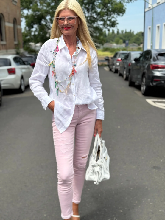 Blondierte Frau mit bunter Perlenkette, weißer Bluse und rosa Skinny-Jeans geht lächelnd mit weißer Tasche auf Asphaltstraße. | © Renate Zott