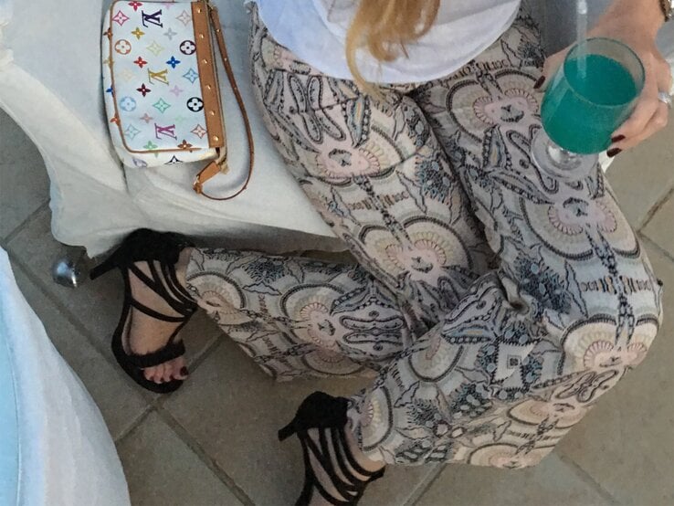 Eine Frau in bedruckter Hose, schwarzen High Heels und weißem Shirt hält ein türkisfarbenes Getränk, daneben eine Designer-Handtasche.