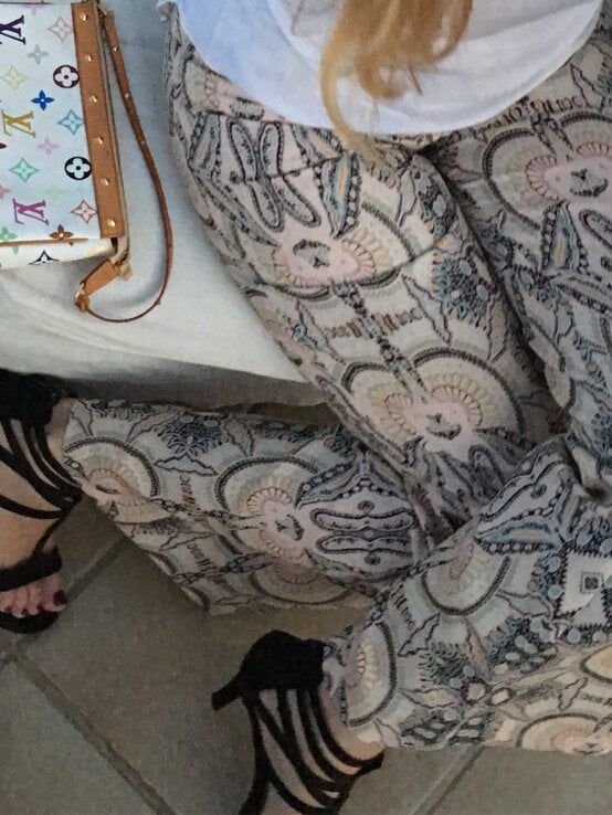 Eine Frau in bedruckter Hose, schwarzen High Heels und weißem Shirt hält ein türkisfarbenes Getränk, daneben eine Designer-Handtasche. | © Renate Zott