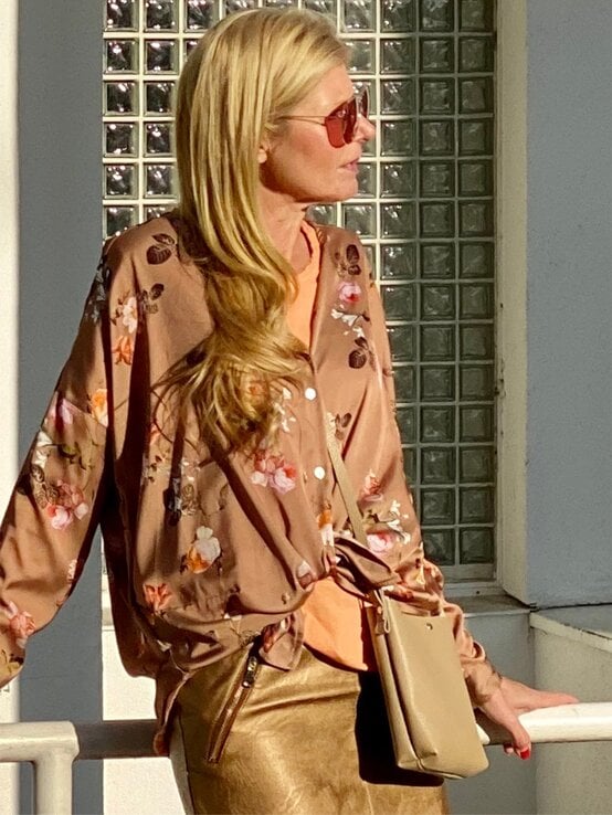 Eine Frau mit langen blonden Haaren trägt eine rosafarbene Bluse mit Blumenmuster, einen goldenen Rock und eine kleine beigefarbene Tasche. | © Renate Zott