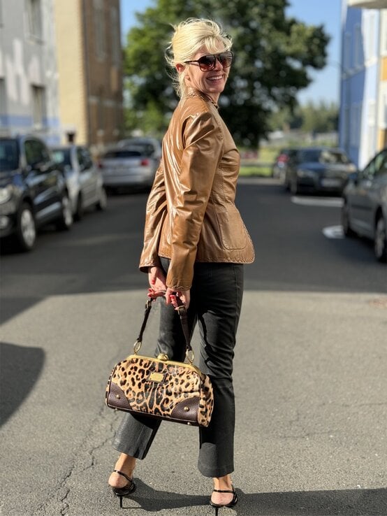 Eine Frau in brauner Lederjacke, schwarzer Hose und Sandaletten trägt eine Leopardenmuster-Handtasche auf einer sonnigen Stadtstraße. | © Renate Zott
