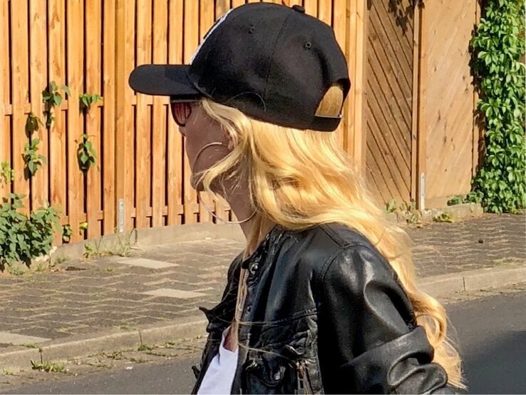 Seitenansicht einer Person mit lässigem Look aus Lederjacke, Cap und Sonnenbrille, umgeben von sommerlicher Szenerie an einem Holzzaun.