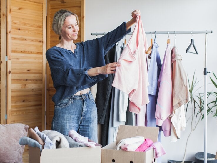 Eine blondhaarige Frau in Jeans und blauem Oberteil hält ein rosafarbenes Shirt vor Kleiderständer und Kartons mit Kleidung hoch.
