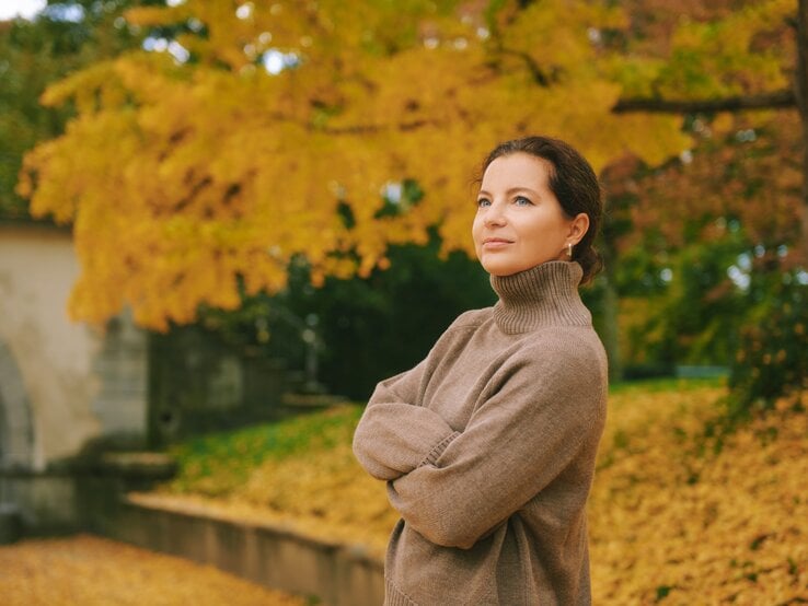 Eine Person in einem hellbraunen Rollkragenpullover steht entspannt vor Herbstbäumen mit goldgelbem Laub und einem steinernen Torbogen.