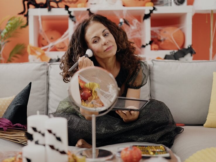 Eine Frau in glitzerndem schwarzen Kleid sitzt auf einem grauen Sofa, mit Halloween-Dekoration und Kürbissen im orangefarbenen Hintergrund.