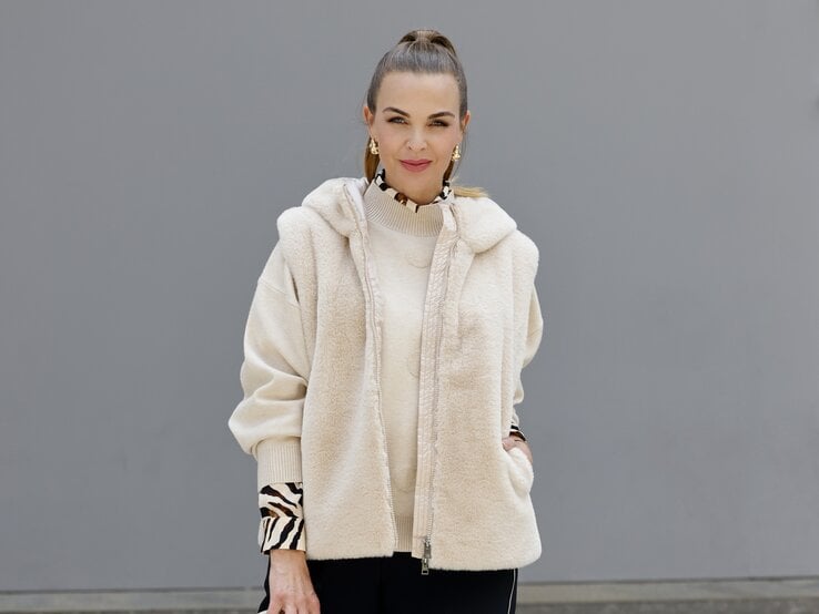 Annika Lau in cremefarbenem Plüschmantel und hellem Rollkragenpullover mit Zebra-Muster-Details steht vor einer grauen Wand. | © Streetstyleshooters for QVC