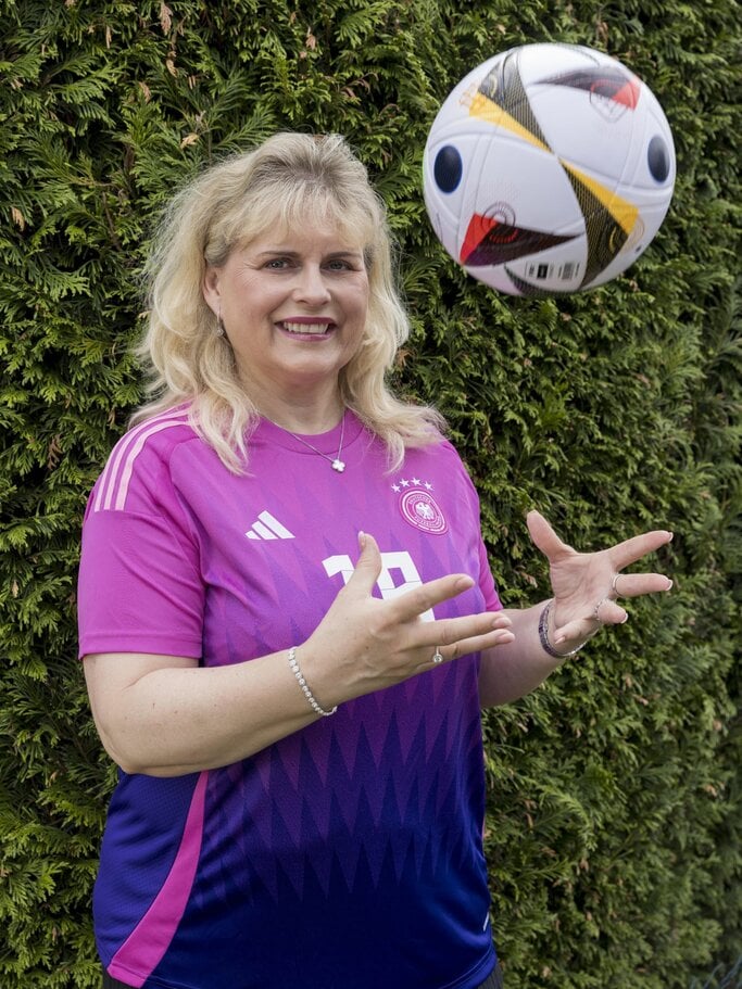 Eine Frau mit schulterlangem blondem Haar trägt ein pinkes Trikot und posiert lächelnd vor einer Deutschlandflagge. | © Anika Büssemeier/ BILD der FRAU