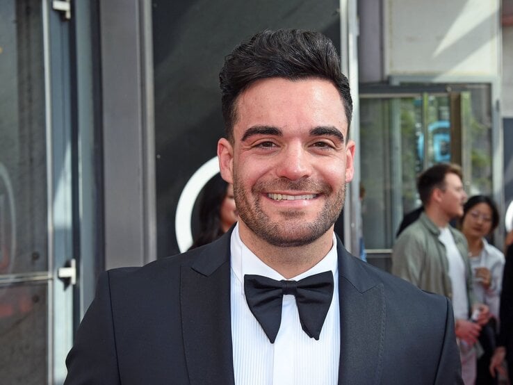 Stefano Zarrella: Ein junger Mann mit dunklen Haaren und Bart lächelt in einem schwarzen Smoking mit Fliege vor einer gläsernen Event-Location.