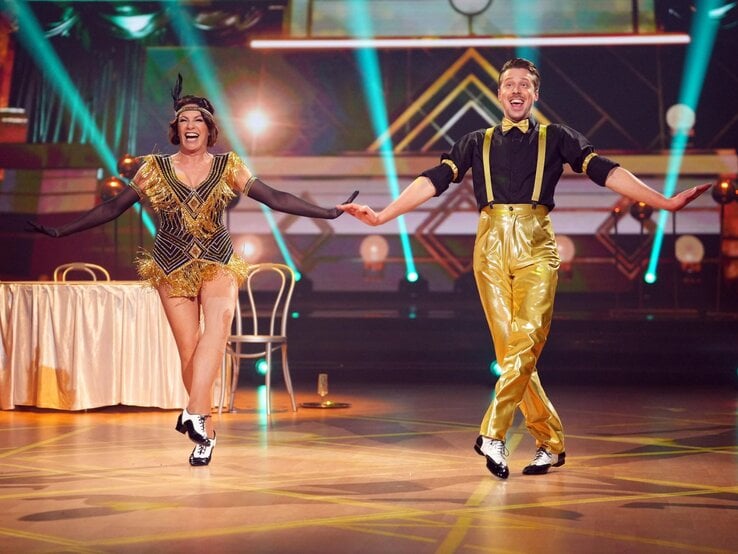Lachendes Tanzpaar (Christine Neubauer & Valentin Lusin) im goldenen Charleston-Outfit tanzt synchron auf beleuchtetem Parkett vor Art-Déco-Kulisse. | © RTL / Stefan Gregorowius