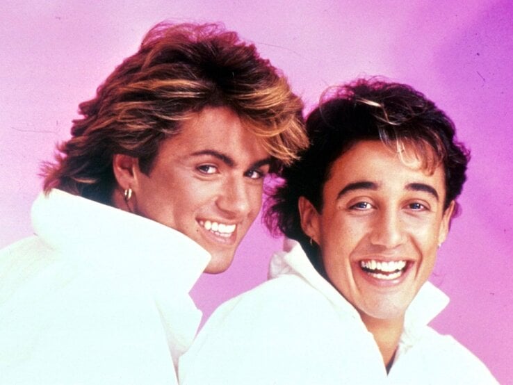 Zwei junge Männer mit Ohrringen und Föhnfrisuren der 80er lachen breit vor rosa Hintergrund in weißen Hemden mit Stehkragen.