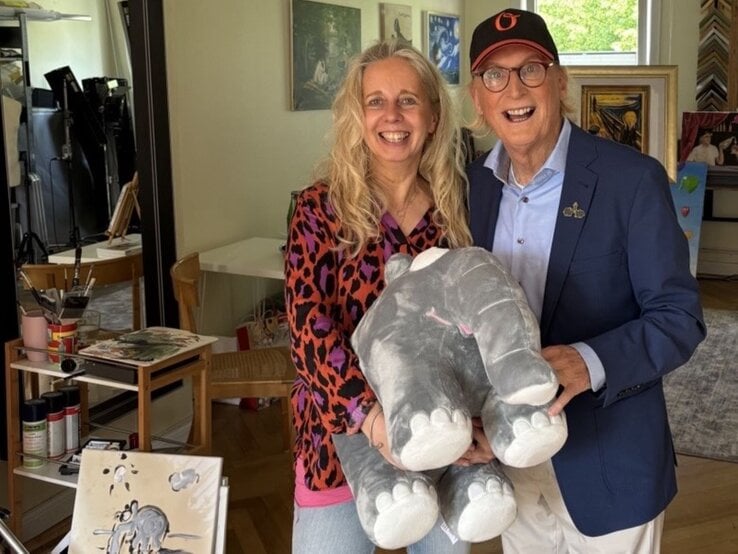 Eine blonde Frau in bunter Bluse und ein älterer Mann mit Brille und Basecap halten lachend ein graues Stoffelefanten-Kuscheltier.