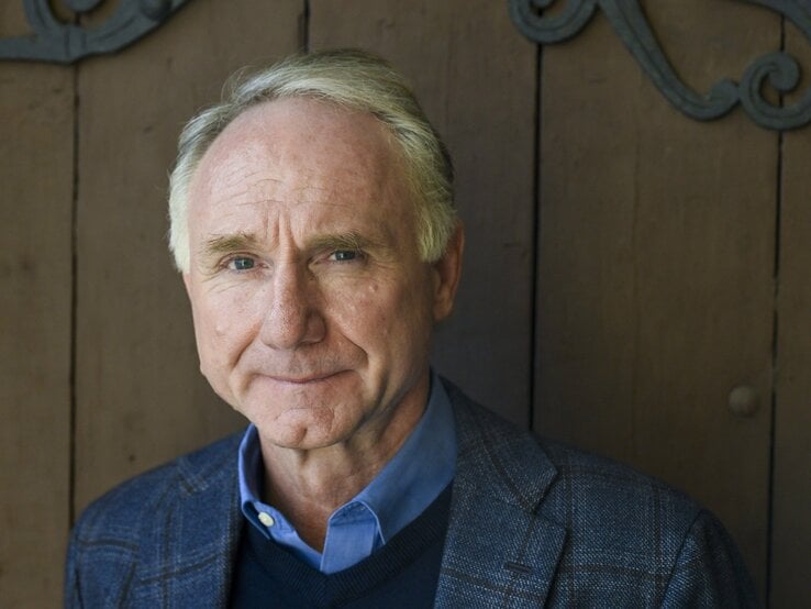 Dan Brown trägt ein dunkles kariertes Sakko mit blauem Hemd und Pullover, im Hintergrund eine schlichte Holzwand mit Metallverzierungen.