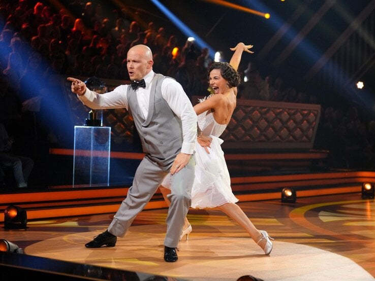Fabian Hambüchen im grauen Anzug tanzt energiegeladen mit lachender Anastasia Maruster in weißem Kleid auf beleuchteter Showbühne.